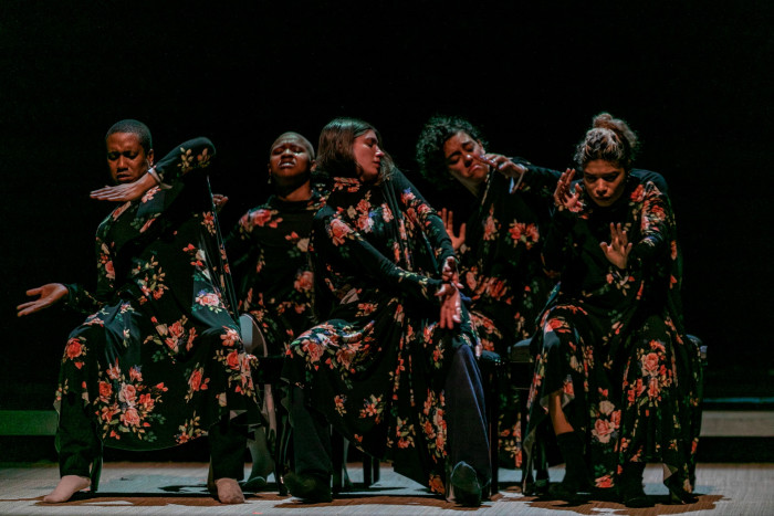 A group of  female dancers wearing dark floral gowns adopt postures and expressions that signify anguish and unhappiness 