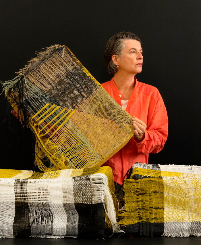 A woman in an orange cotton top, looking severe, holds a panel of yellow and black yarn