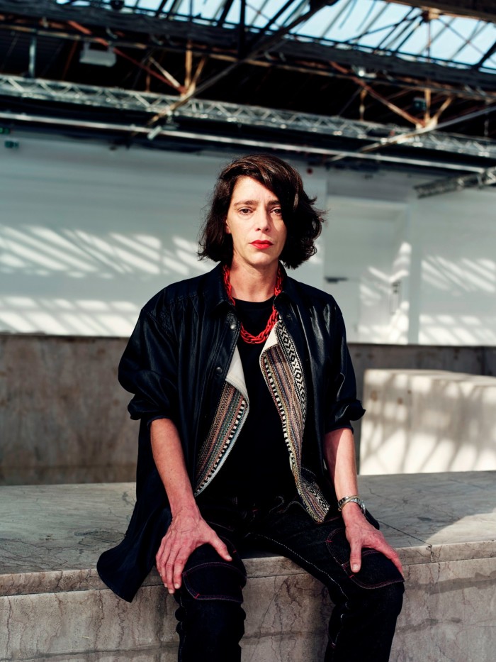 A woman in a black jacket and black trousers with red lipstick and a red necklace looks severe as she sits on a concrete bench