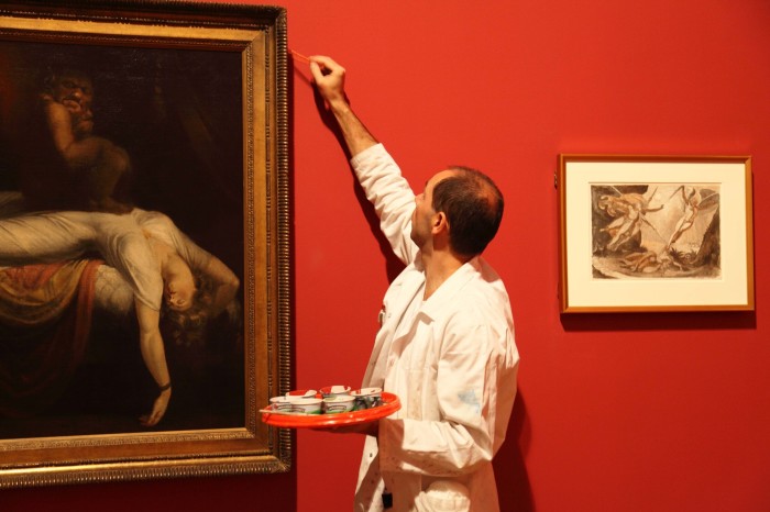 A man in a white jacket dabs at a red wall next to an oil painting with red paint
