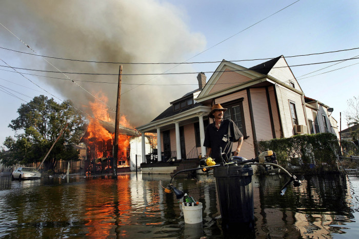 A man stands in floodwater on a street while a house burns down behind him