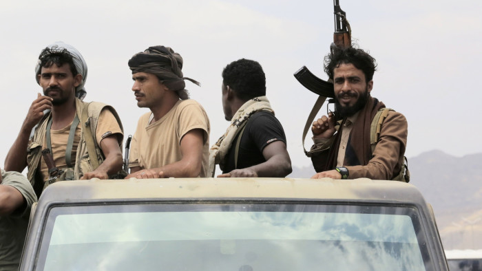 Four men, one holding a rifle up, ride in the back of a pick-up truck