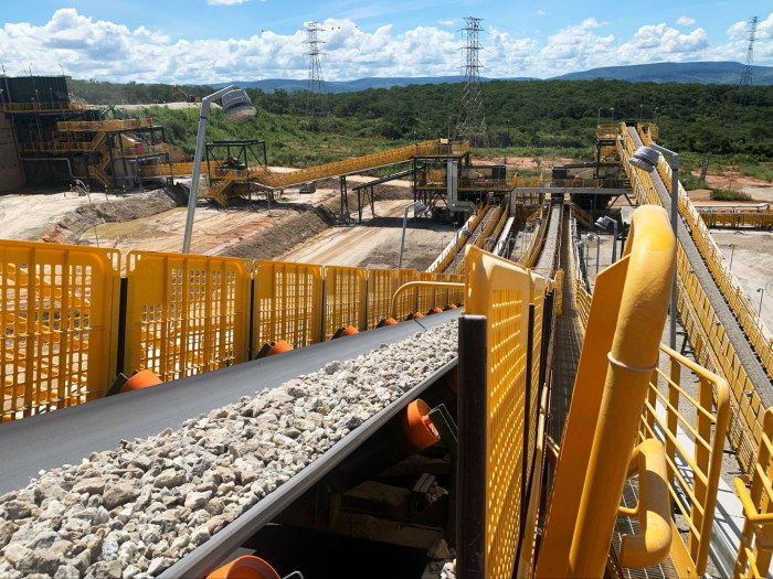 A conveyor belt loaded with gravel running through a large industrial mining site with various machinery and structures in the background, all secured by bright yellow safety barriers