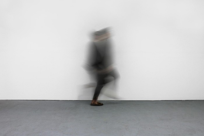 The blurred silhouette of a person dressed in dark clothes stands in motion in the centre of an empty room