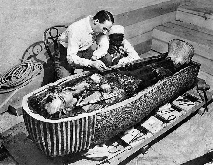 A man in a white shirt poking into an open coffin, complete with mummy