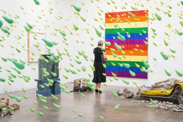 A woman views a rainbow-coloured rectangular art work on a wall while green flecks of plastic in the shape of raindrops hand from the ceiling behind her
