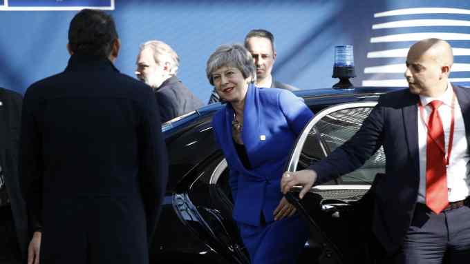 Theresa May, smiling and wearing a blue suit, gets out of a car as a man in a suit and red tie holds the door open for her