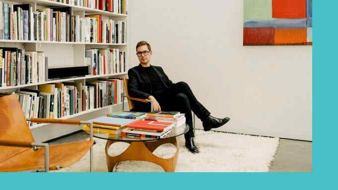 A man all in black reclining in a chair in front of a wall full of art books