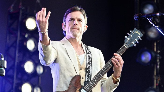 A man with shortish black hair in a cream jacket holds a guitar in one hand and raises his other