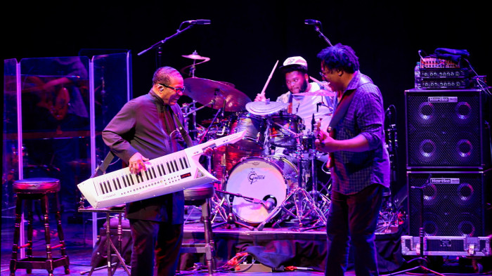 A man stands on stage and plays a keytar with a drummer and bass player