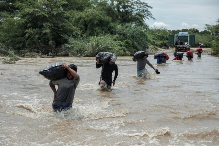 A line of people waist high in water carrying sacks of vegetables across a fast-moving tropical river