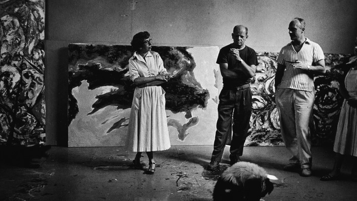 A woman and two men stand together in conversation in an artist’s studio, with large abstract paintings propped up on a wall behind them