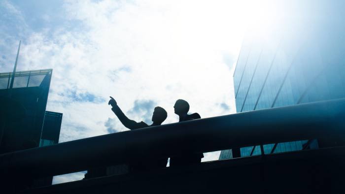 Two men are silhouetted against the concrete rails of what appears to be a bridge against a background of a building on either side and a clear sky