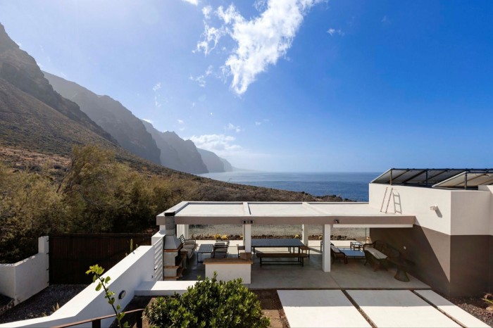 The view from the side of a villa, looking through its sheltered terrace towards a rugged mountainous coastline under a blue sky