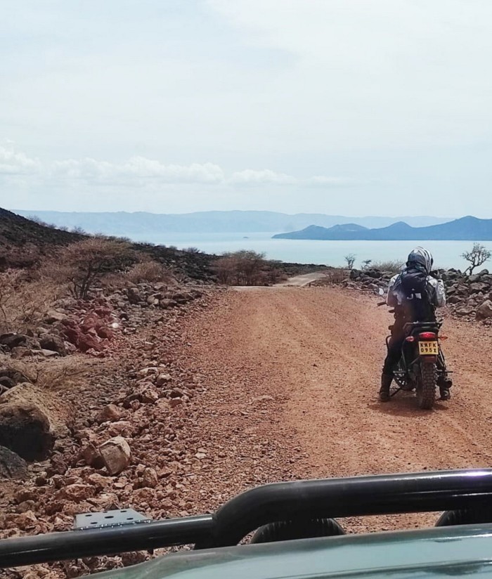 A woman on a motorbike driving down a sandy road towards a lake in the distance