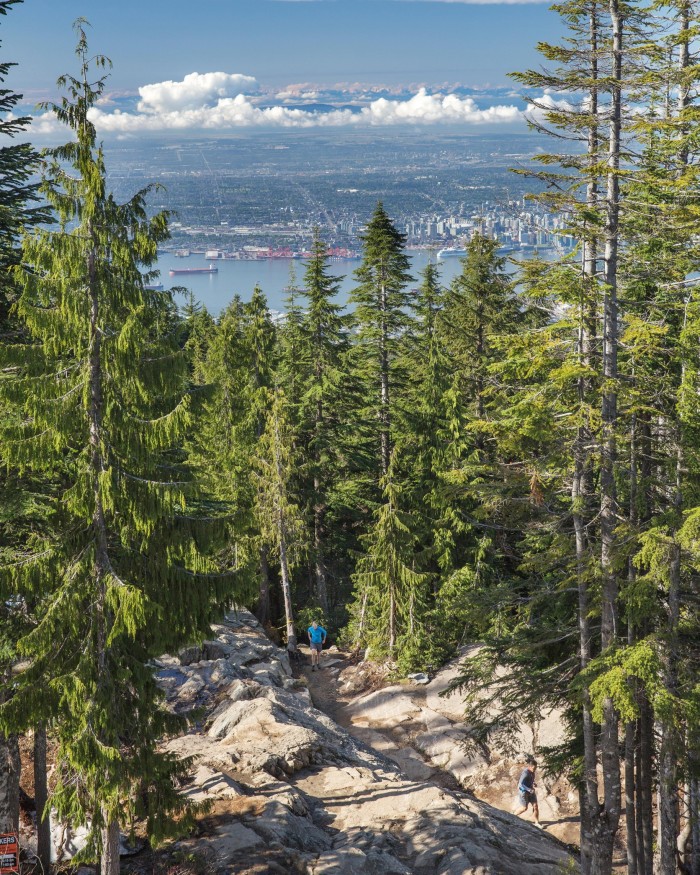 A man running up a tree-flanked steep rocky incline on the Grouse Grind, with the bay and Vancouver’s skyline in the distance behind him