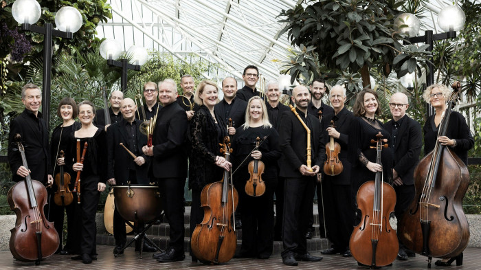 A group of about 20 people holding instruments and wearing black smile in a large leafy conservatory