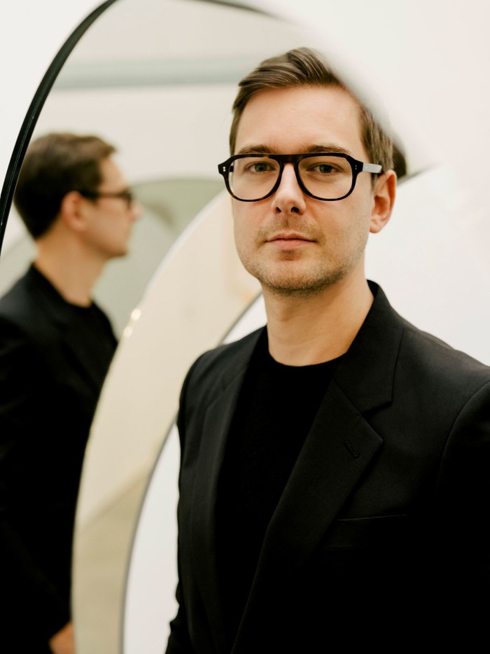 Photo of a youngish man with stubble in thick black glasses looking warily at the camera