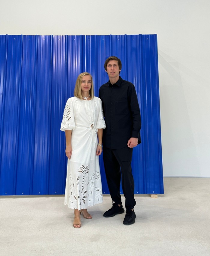 A woman in white and a man in black stand in front of a large blue sculpture
