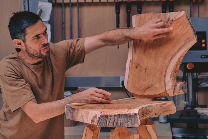 A man in a brown T-shirt is touching a chair made from irregular chunks of wood