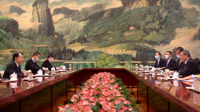A high-level diplomatic meeting unfolds in a room decorated with an elaborate mural of a mountainous landscape. Officials from both sides sit at a polished wooden table, separated by an arrangement of pink flowers, while some attendees wear facemasks