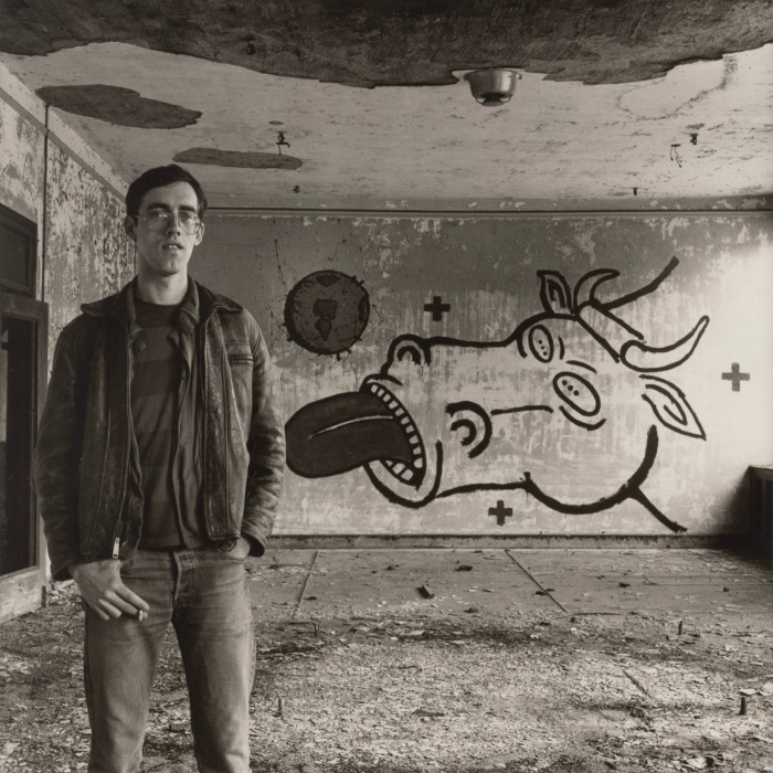 Black and white photo of a man in jeans standing in a semi-abandoned room with a mural of a cow sticking its tongue out