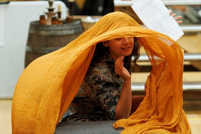 A woman leans on a wooden crate as an orange silk scarf cascades over her head