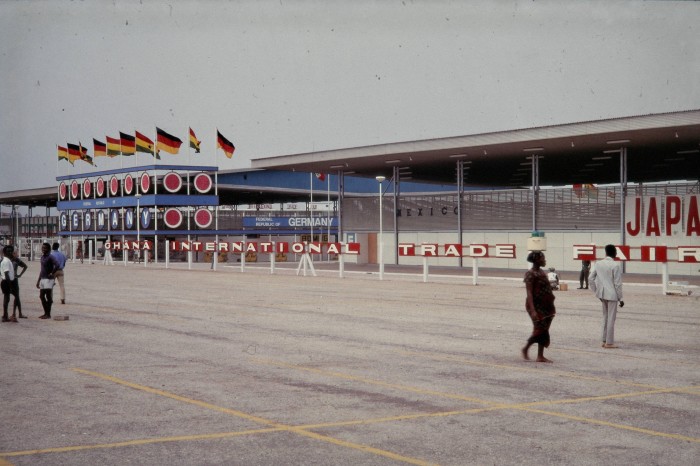 Rows of flags in front of a large modernist building with people walking in front of it 