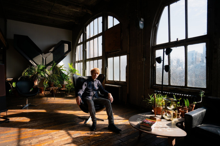 A man in a shirt and jeans sitting in a modern leather chair in a loft studio with plants on a palette-shaped table