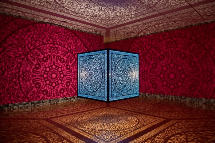 A bright blue-glass box covered in an intricate design illuminated from inside casts patterned shadows on red walls