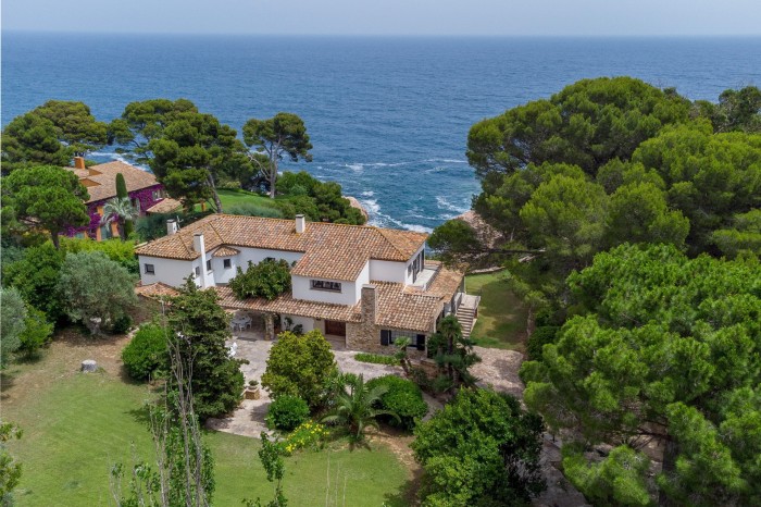 An overhead view of a whitewashed home with terracotta tiles surrounded by trees and facing the sea 
