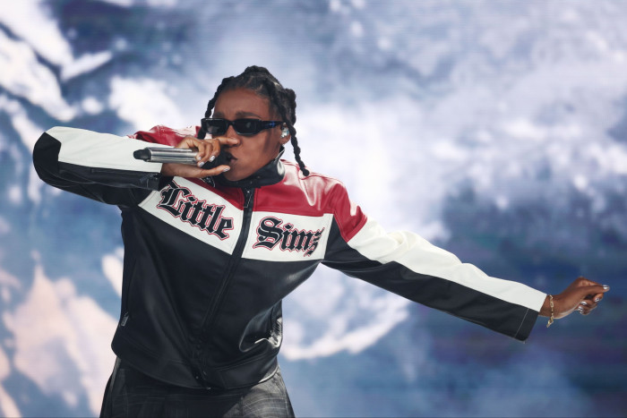 A woman wearing a motorcycle jacket saying “Little Simz” and sunglasses raps into a microphone