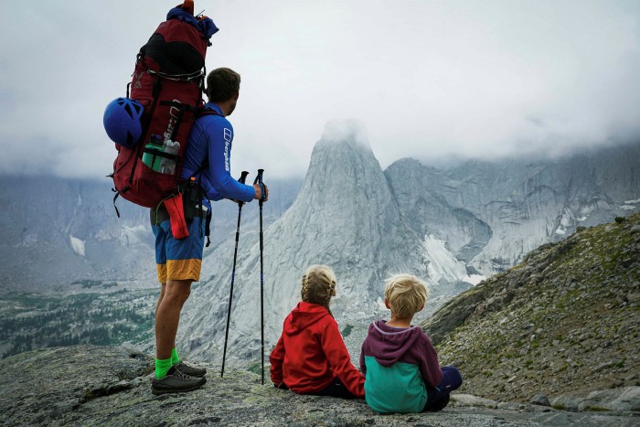 Leo Houlding, wearing his backpack, stands looking towards a mist-covered peak. Freya and Jackson sit on the ground next to him