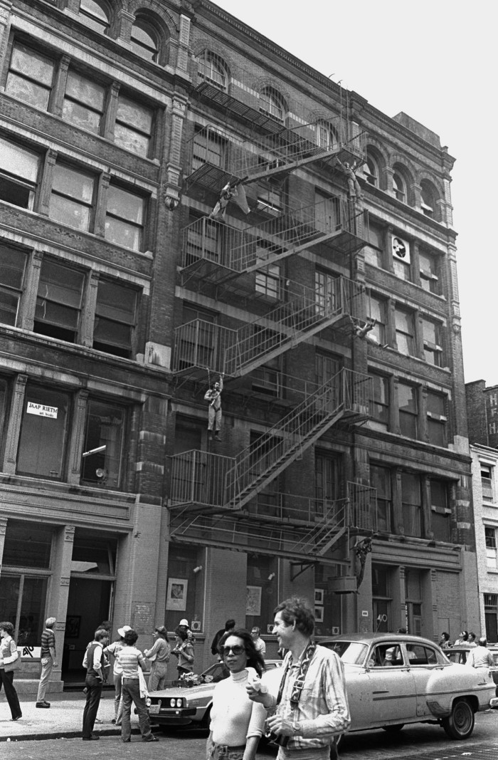 Two artists dangle from a fire escape steps on a New York cast-iron building as a crowd in the street looks up at them