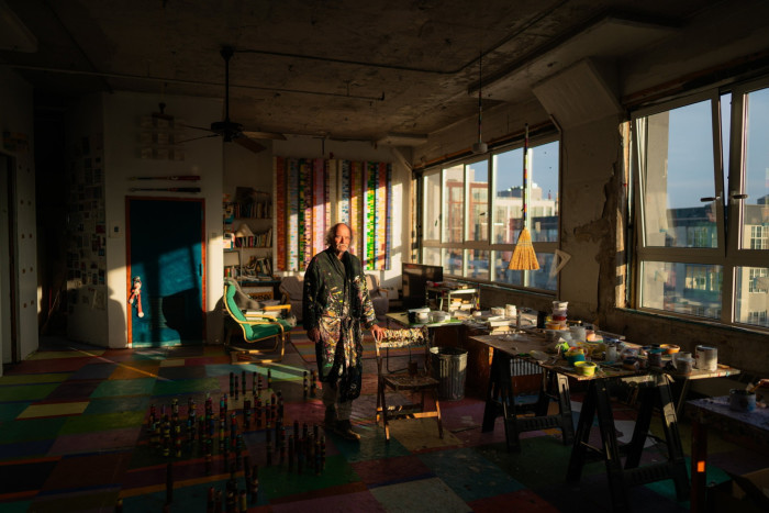 A man in paint-spattered overalls stands in a loft studio as sunlight streams in through the windows
