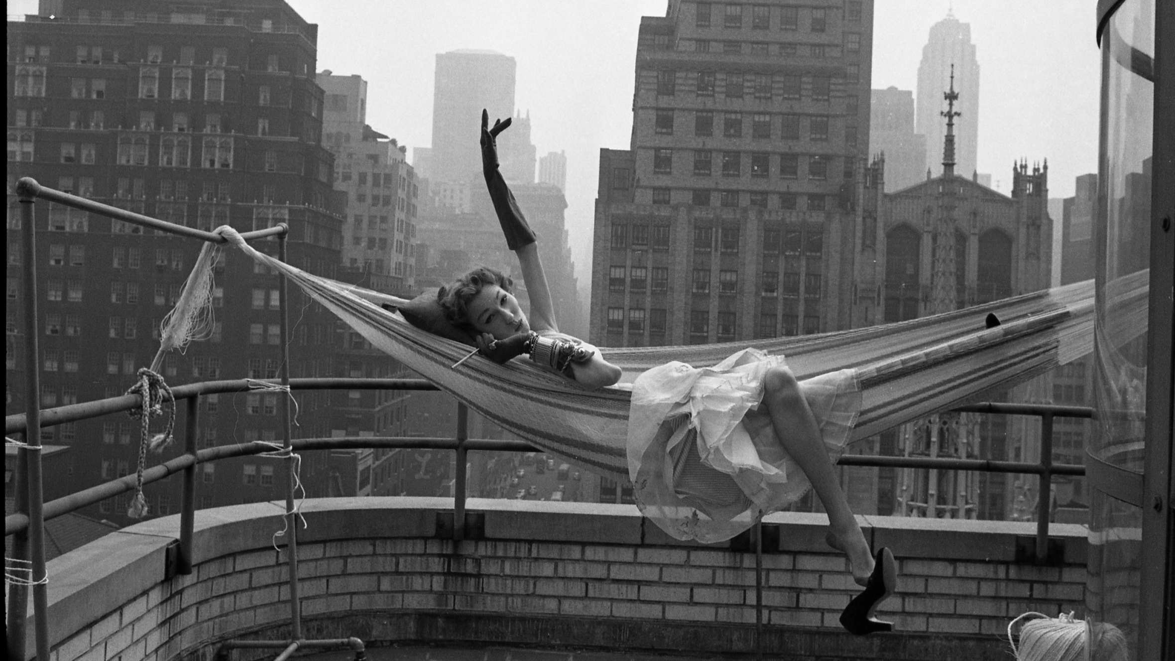 A glamorously dressed woman lies languidly in a hammock on the rooftop of a building surrounded by skyscrapers