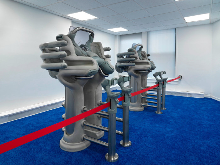 In a bright office space with blue velvet flooring, two grey totemic sculptures featuring alien-like hand components stand before a red belt barrier