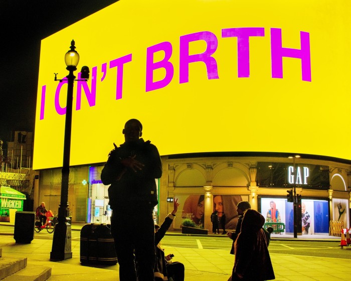 In a large public plaza, a wide screen on top of a building reads ‘I CN’T BRTH’ in bright pink against a yellow background while a young man dressed in black poses in front of it