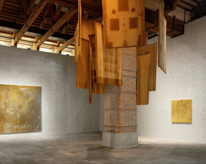 Two golden-brownish-yellowish canvases hang on adjacent walls near a display of golden-orange fabric banners hanging from the ceiling
