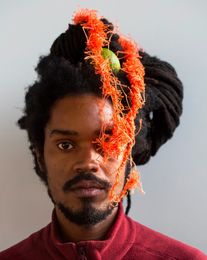 A man stares straight at the camera. His hair is covered in a sort of orange braid