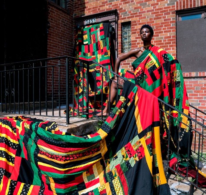 Photo of a proudly posed woman draped in a red, yellow, green and black patchwork textile which descends from her to the ground and swathes everything