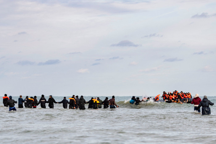 A lot of people in a small rubber boat wearing orange life vests. There is a line of people holding hands from the shore to the boat. Some people are struggling in the waves