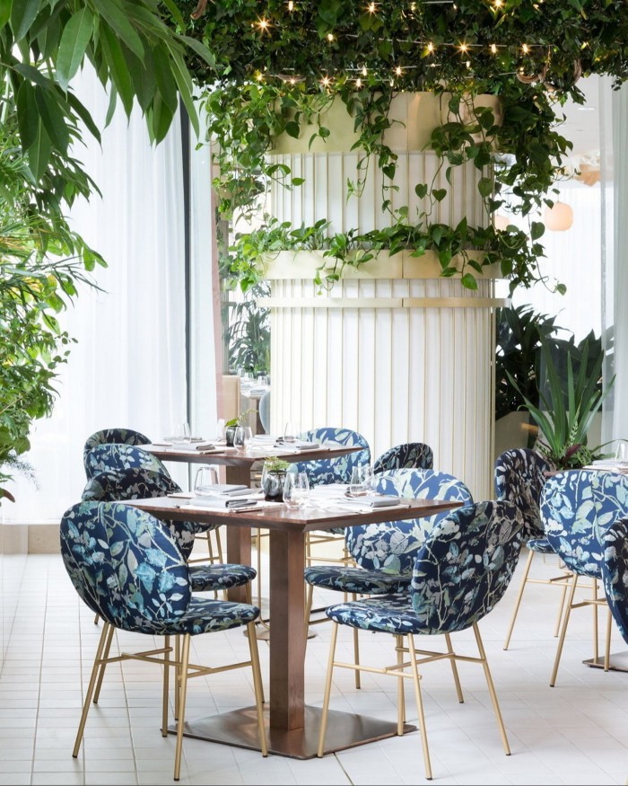 Blue floral-patterned chairs around brass tables in the Botanist restaurant, with plants winding around large floor-to-ceiling white pillar