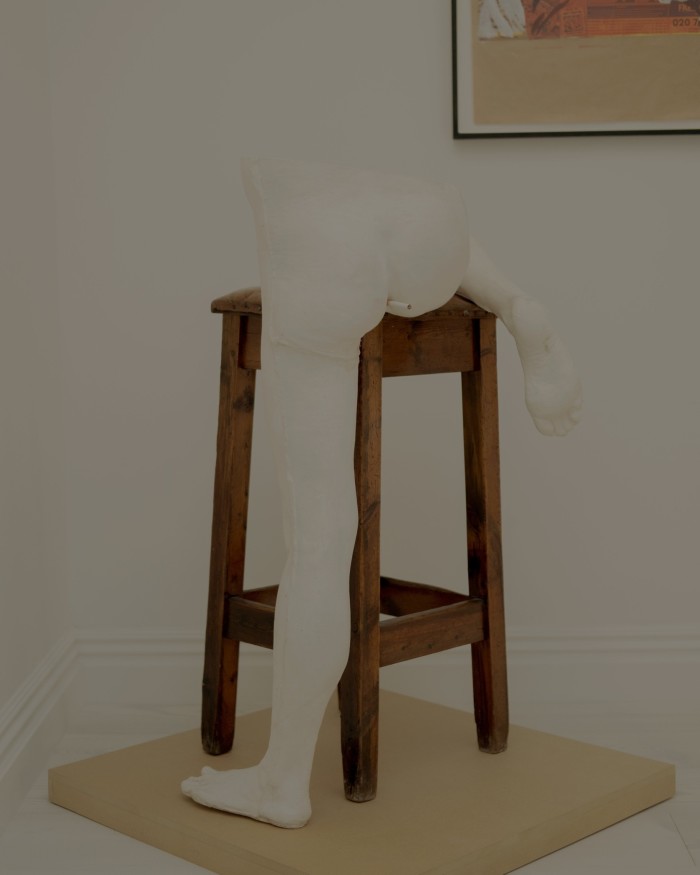 Sculpture of a naked person from the waist down, their legs over a bar stool, a cigarette stuck in their bottom