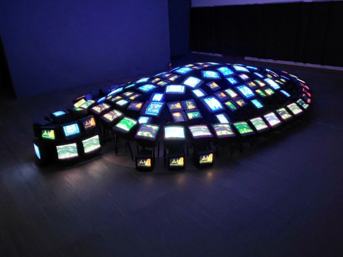 A series of glowing television monitors arranged on the floor of a gallery to resemble a turtle