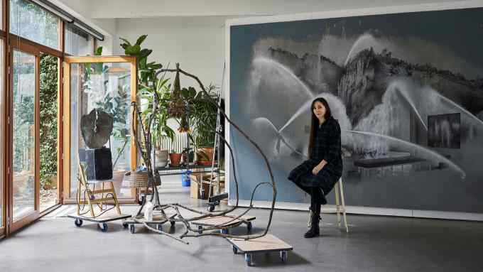 A woman sits on a high stool in front of a large grey painting next to a skeletal sculptural assemblage