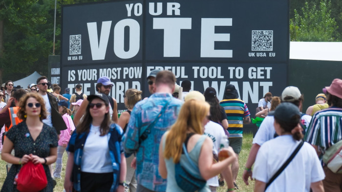 Festival goers walk through a field beneath a giant banner calling for people to vote