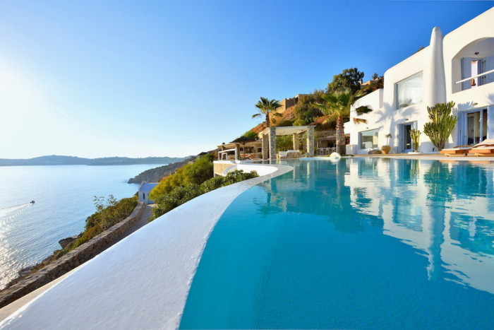 A hillside white Greek villa reflected in an infinity pool overlooking the sea