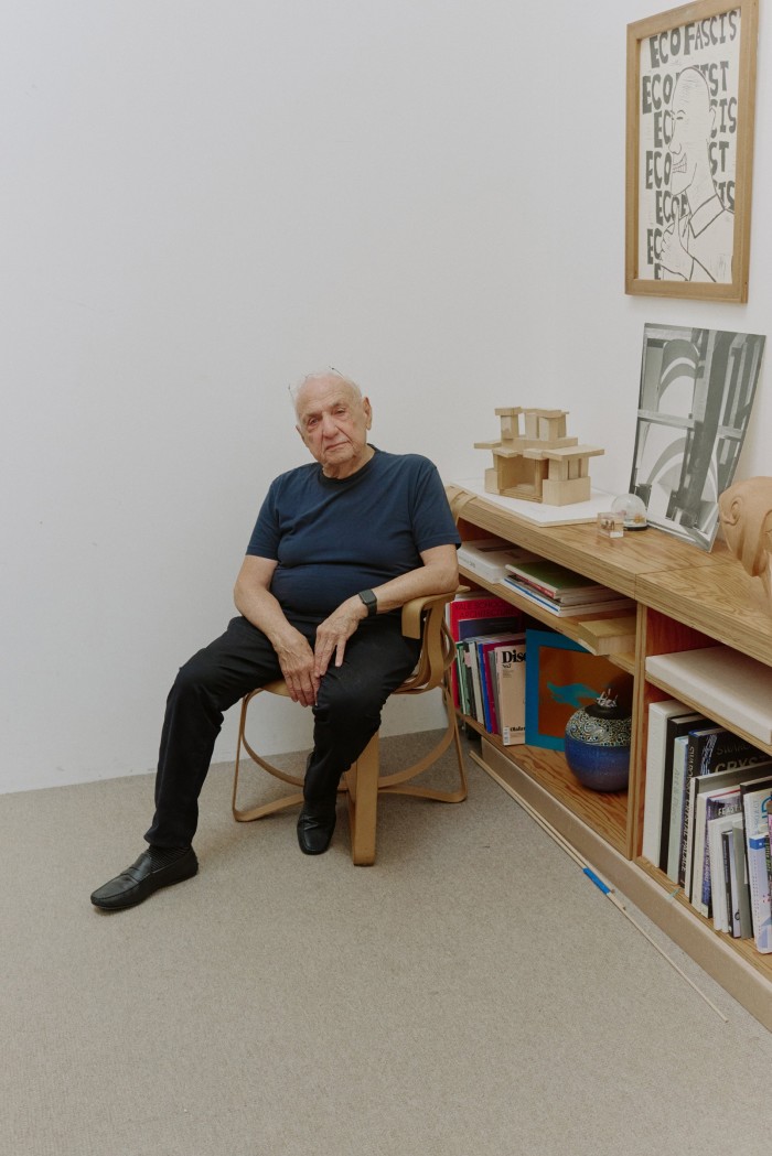 Gehry sits in the studio next to a bookshelf