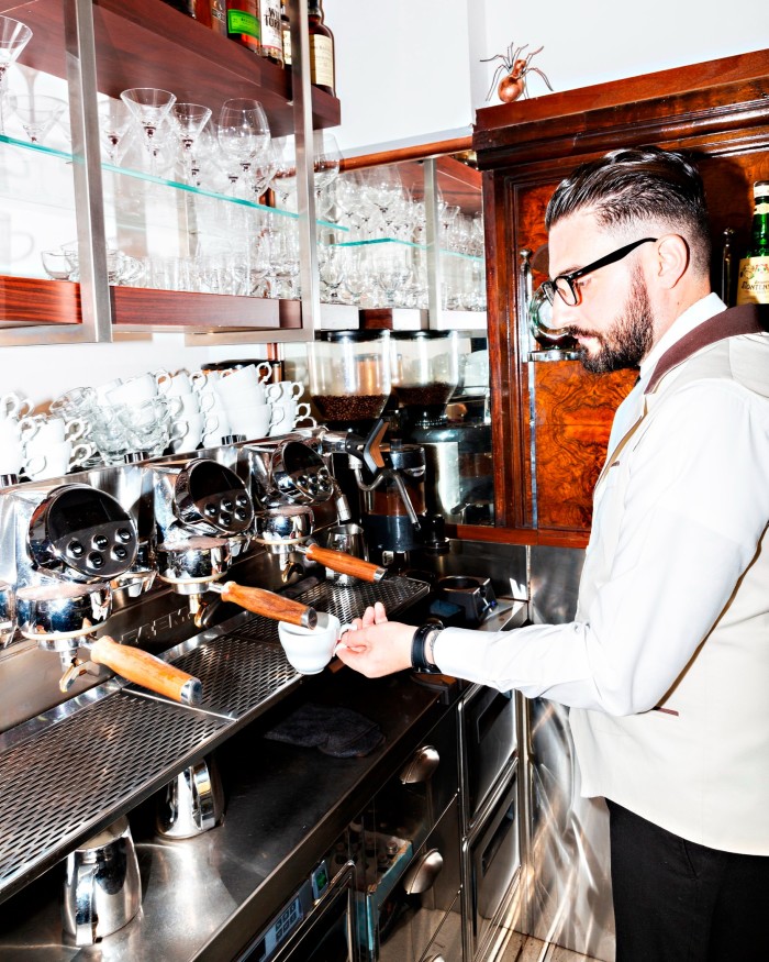 A bearded and bespectacled young barista in a pale-brown blazer and black trousers preparing a cup of coffee at a machine behind the counter in Taveggio